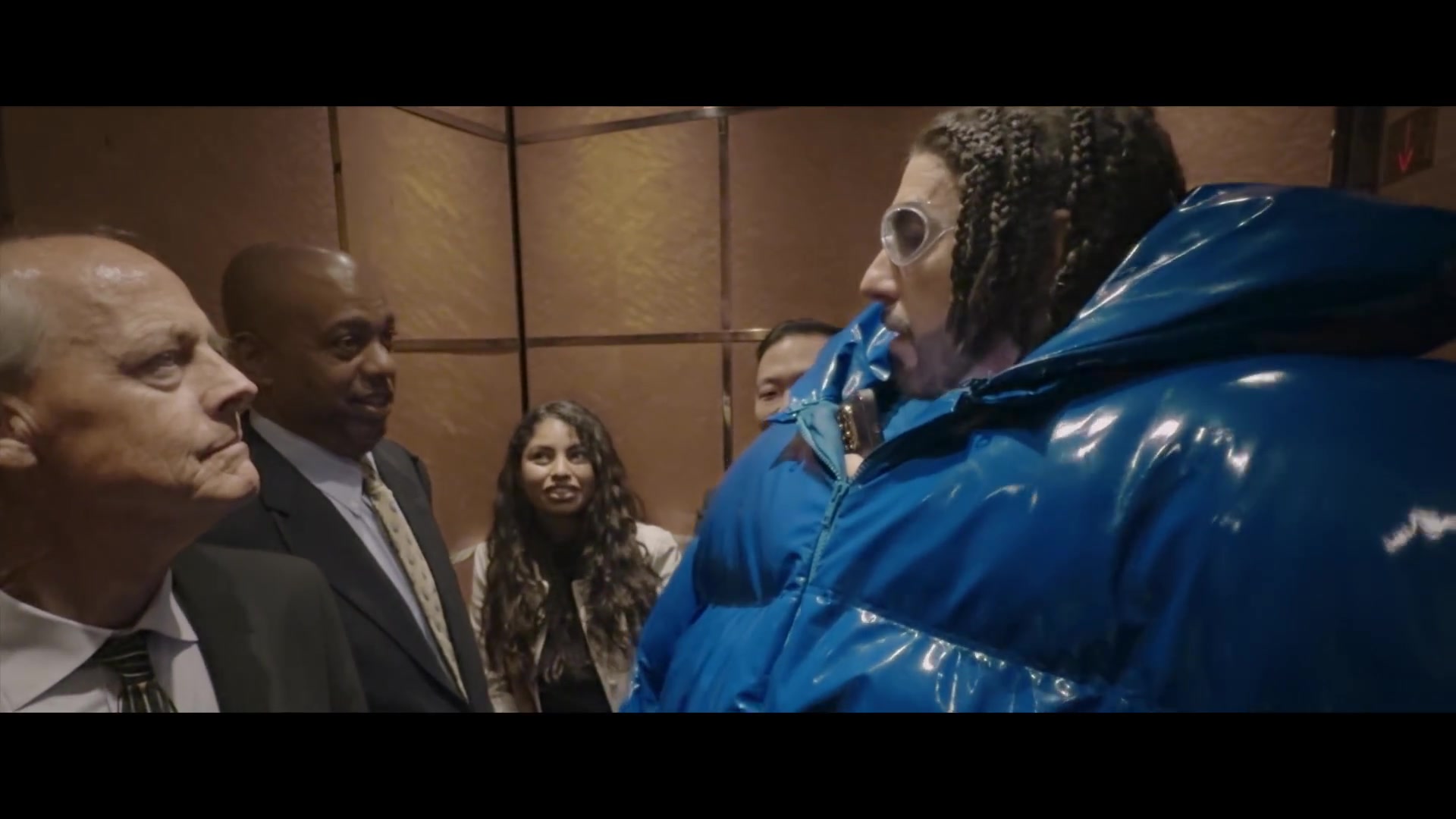 Ali G in een blauwe glimmende jas in een lift vol mannen in pak, uit de trailer van Ali G: Who Iz I?