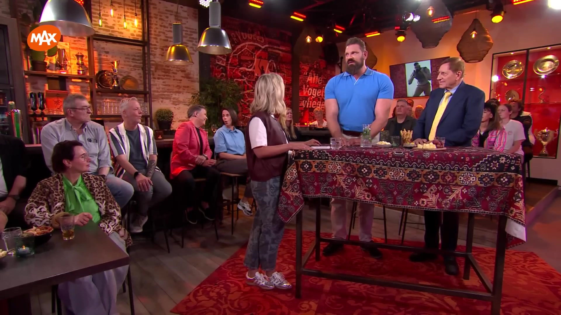 Carrie ten Napel in de studio van Carrie op vrijdag met gasten en publiek rond een tafel met een tapijtkleed