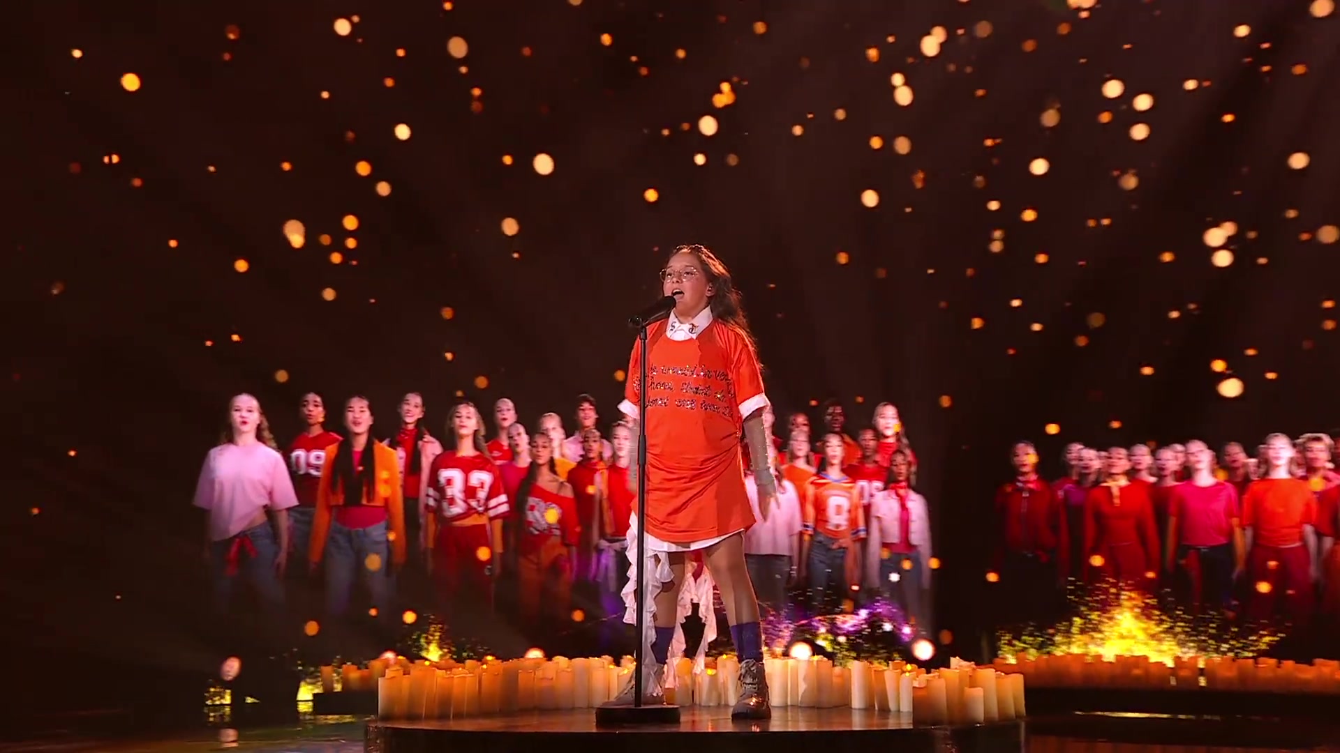 Maria zingt Stronger Together in een oranje jurk met een koor achter zich op het podium op Malta