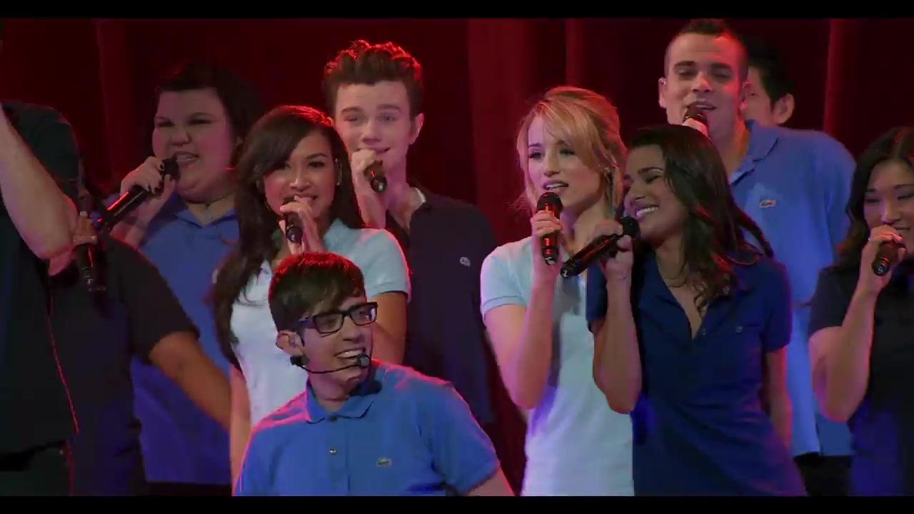 De cast van Glee zingt met microfoons op het podium, met Lea Michele rechts