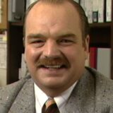 Acteur Richard Riehle met snor en grijs colbert in een interview uit 1999