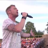 Ronan Keating zingt met een microfoon op een openluchtpodium voor een groot publiek