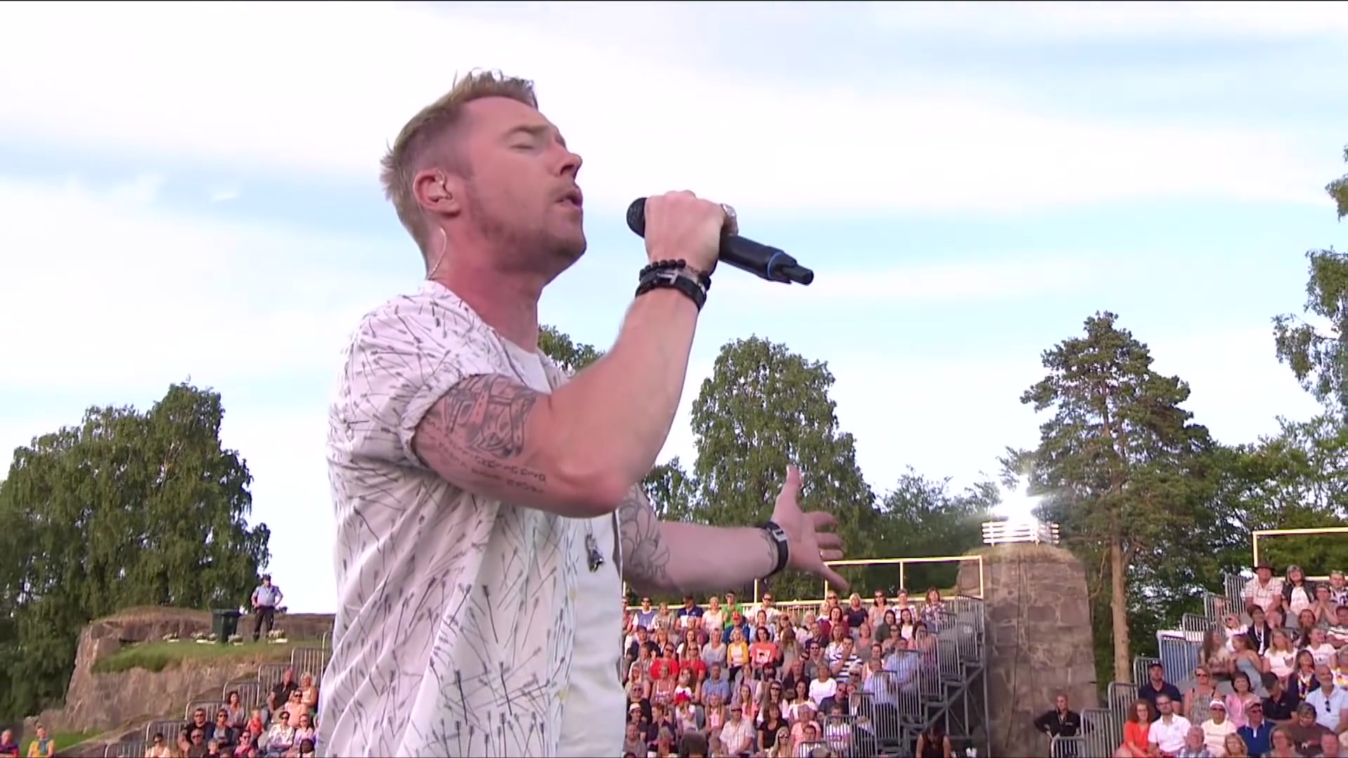 Ronan Keating zingt met een microfoon op een openluchtpodium voor een groot publiek
