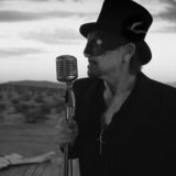Bono met hoge hoed en masker achter een microfoon in de woestijn, uit de videoclip van The Greatest Show On Earth
