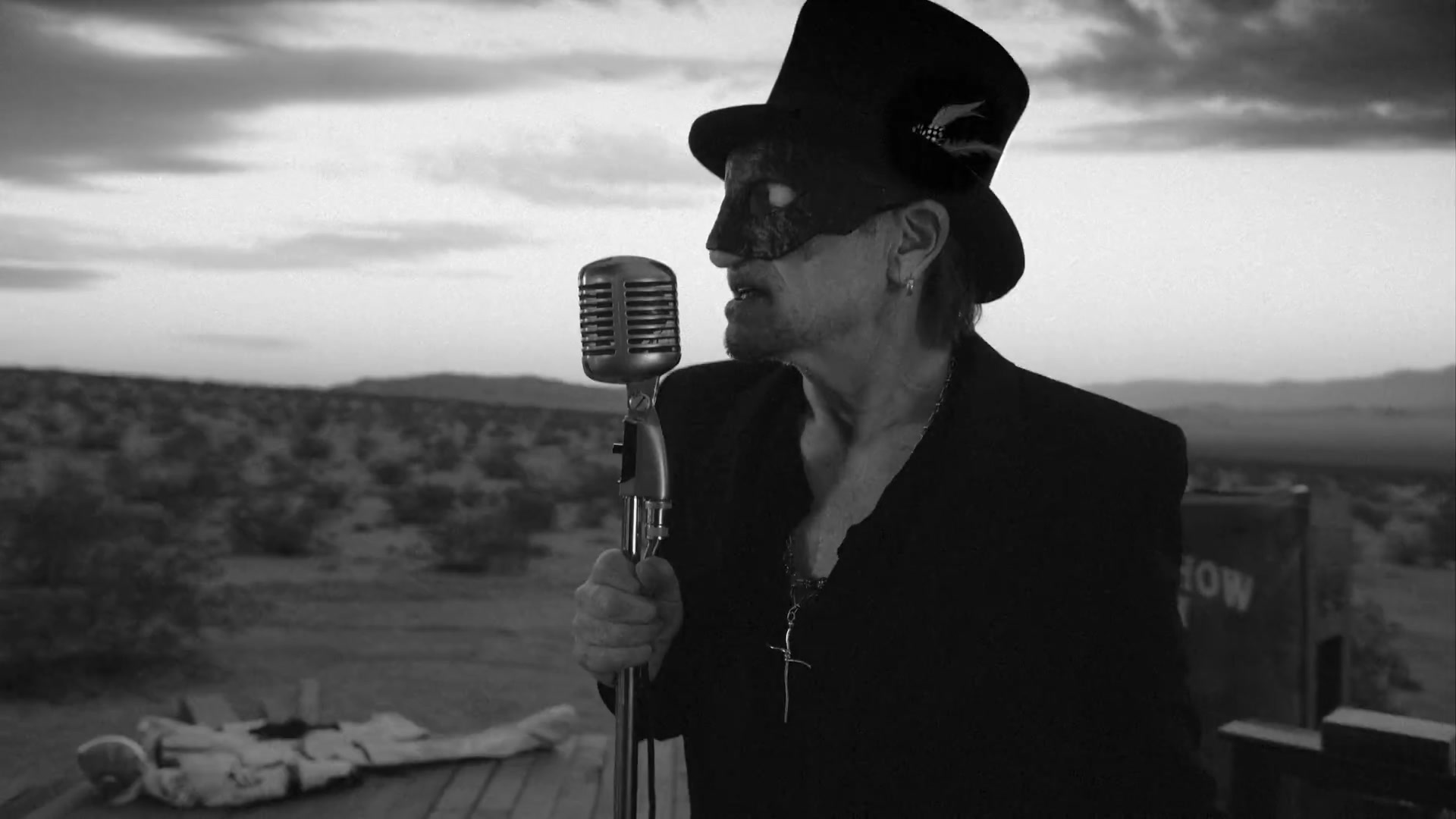 Bono met hoge hoed en masker achter een microfoon in de woestijn, uit de videoclip van The Greatest Show On Earth